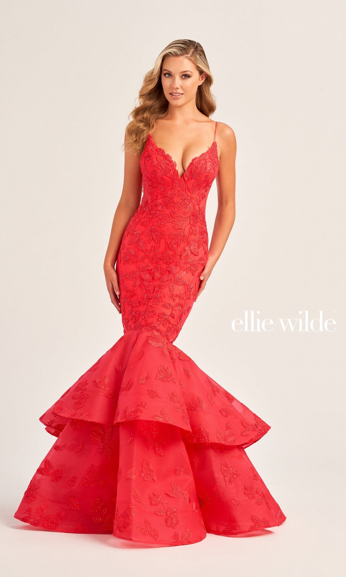 A woman wears the Ellie Wilde Long Lace Mermaid Prom Dress EW35092—fitted, red, with thin straps, a deep V-neckline, and a tiered ruffled skirt. The text